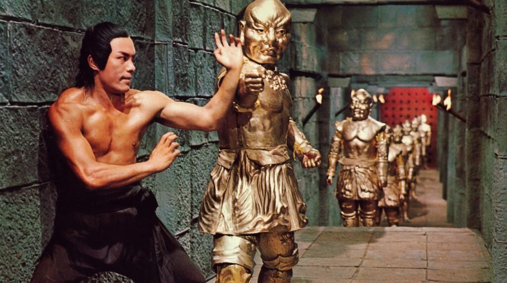 A martial artist in black pants fights a line of golden, armored warrior statues inside a stone corridor lit by torches.