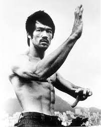 Black and white photo of Bruce Lee shirtless in a martial arts stance, with his arm extended and intense expression, showcasing his muscular physique.
