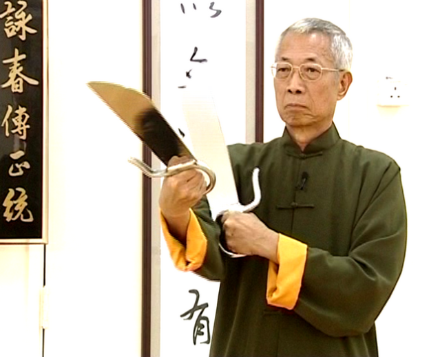 " Wing Chun and MMA Chu Shong Tin, martial arts master wearing a dark green traditional uniform with yellow cuffs, holding a pair of Wing Chun butterfly swords in a training hall decorated with Chinese calligraphy scrolls.