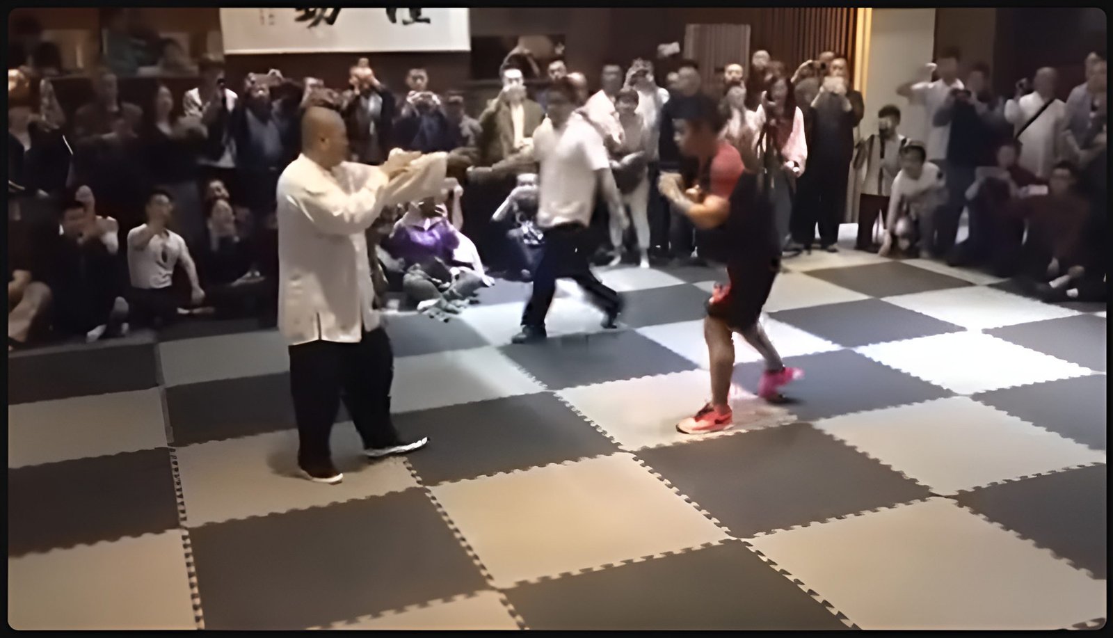 Wing chun vs mma A martial arts match between a man in traditional white kung fu attire and another in modern MMA gear, standing on padded mats surrounded by a crowd of spectators taking photos and watching closely.